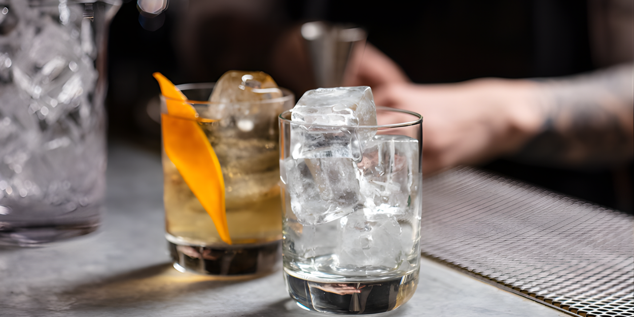 Cocktail with ice and an orange peel next to a glass of water on a bar.