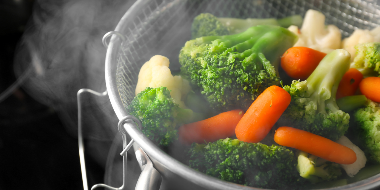 A professional stainless steel steaming basket filled with vibrant broccoli, cauliflower, and carrots.