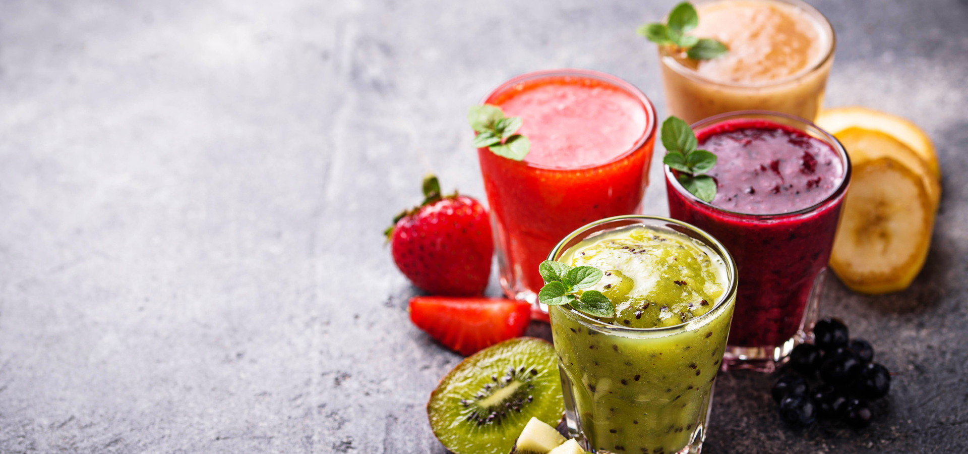 Four colorful smoothies in glasses with fresh fruits on a gray surface
