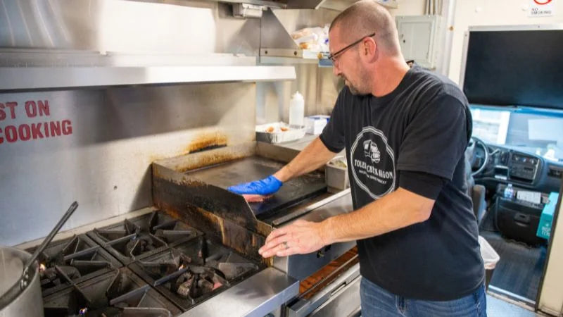 Person cleaning a commercial kitchen stove with a sign in the background.