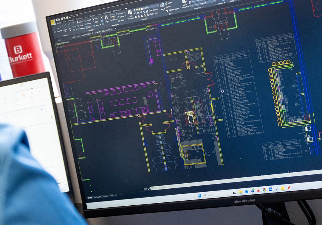 Computer screen displaying technical drawings with a blurred person wearing a blue shirt in the foreground.