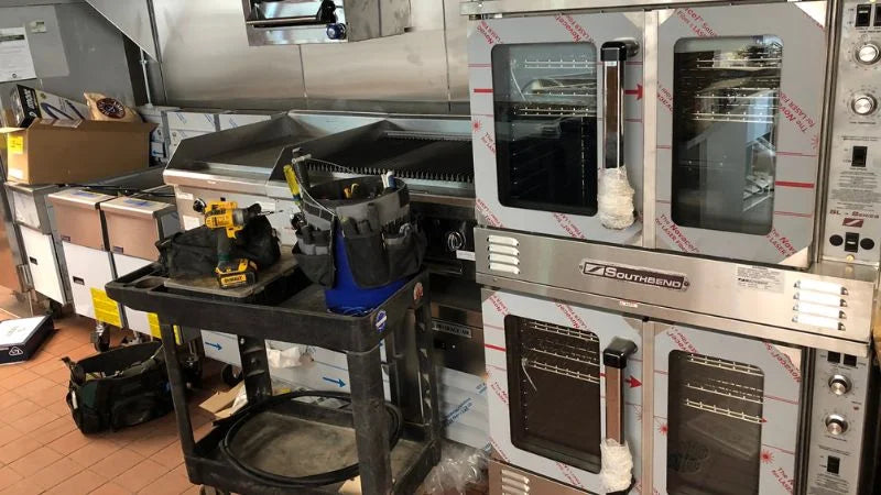 Stacked convection ovens and charbroiler in a commercial kitchen, with a utility cart holding tools and equipment during installation