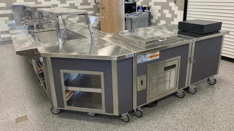 Mobile stainless steel serving station with sneeze guard, refrigerated compartments, and storage on wheels in a cafeteria setting