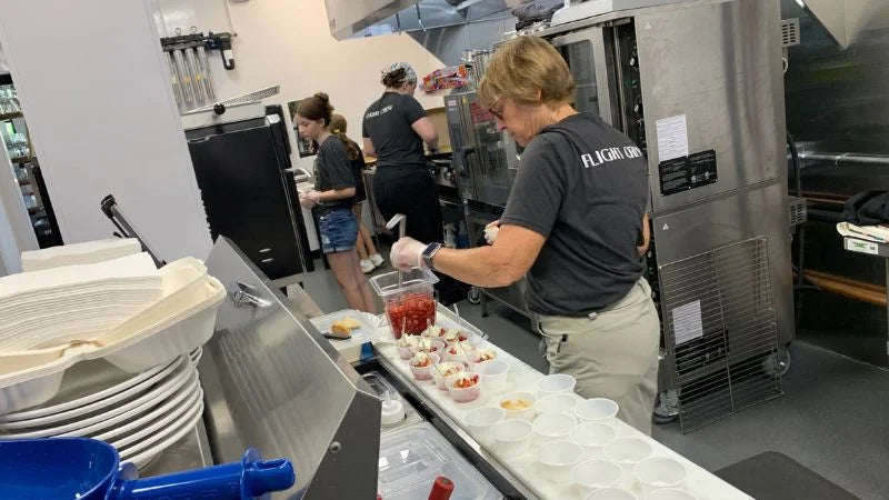 Kitchen staff assembling food portions at a prep line with cups, ingredients, and commercial ovens in the background