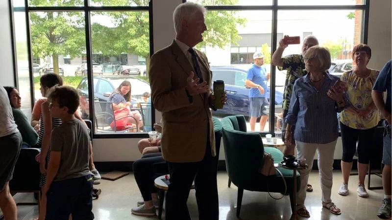 Man speaking to a small group inside a café, with people gathered around and others visible through large windows outside