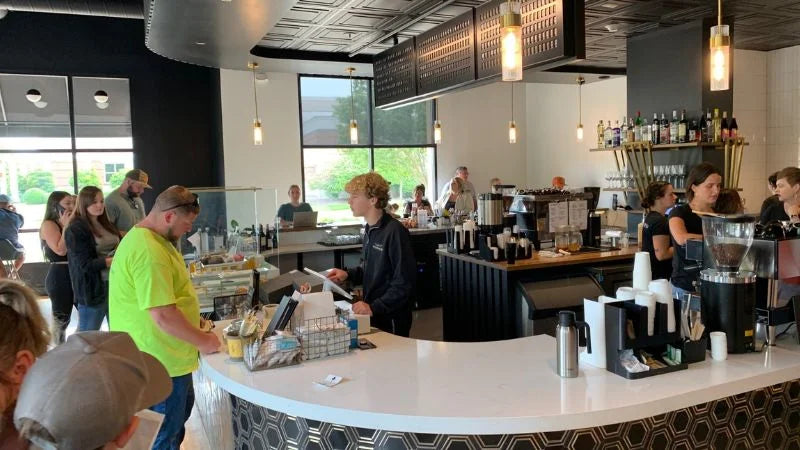 Busy café interior with customers ordering at a curved counter, barista station, and coffee equipment under modern pendant lighting