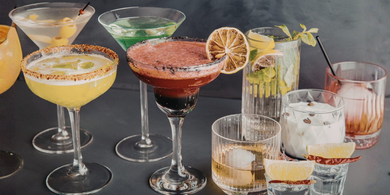 A professional collection of premium beverageware, including elegant cocktail coupes, rocks glasses, and highball tumblers filled with colorful craft cocktails and mixed drinks.