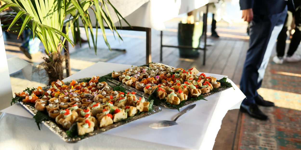 Professional catering display featuring elegant silver trays of gourmet appetizers on a white linen tablecloth during an outdoor event.