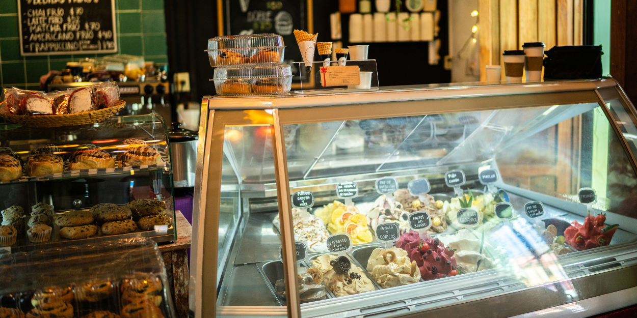 Professional ice cream shop display case featuring assorted gelato flavors in stainless steel pans, with clear protective glass and commercial dessert service equipment.