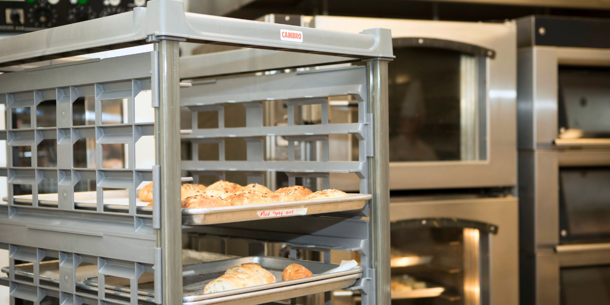 A heavy-duty commercial bun pan rack holding several sheet pans of freshly baked pastries in front of a high-capacity industrial oven.