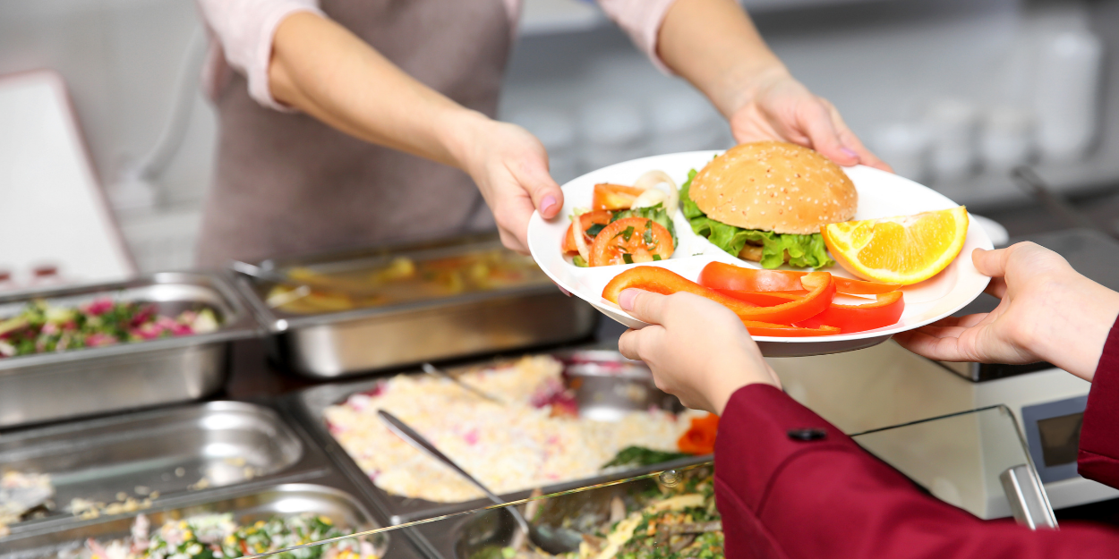 Commercial cafeteria serving line featuring stainless steel food pans and a student receiving a multi-compartment portion control tray with a balanced meal