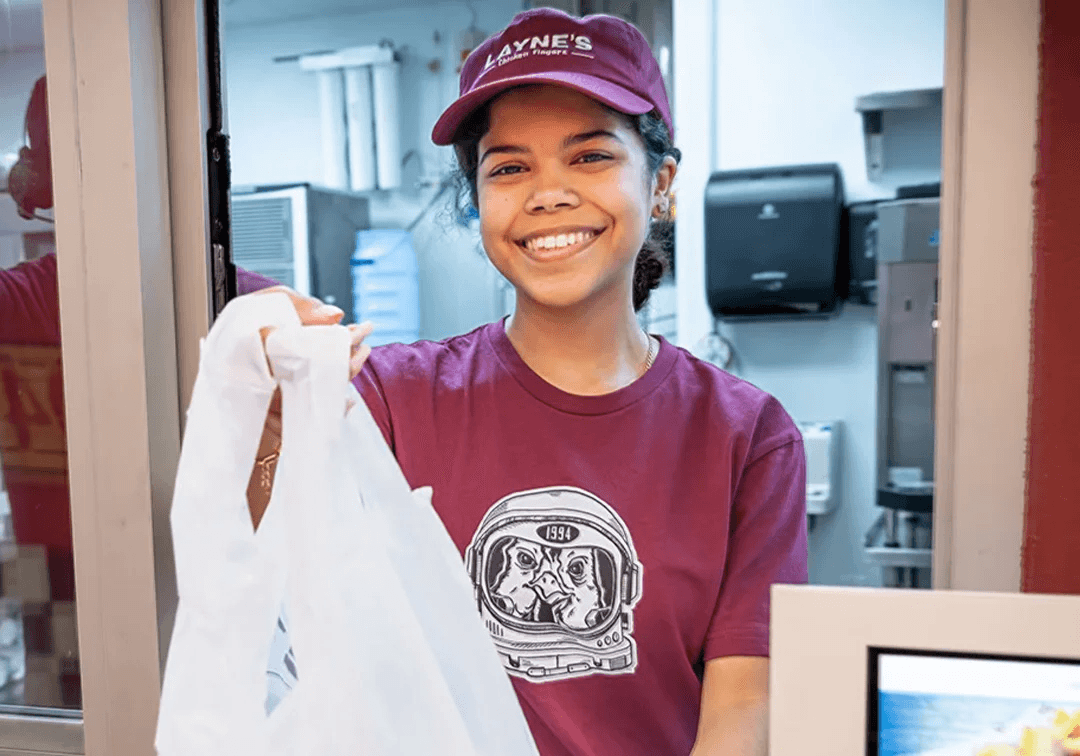Person wearing a purple 'Layne's' cap and t-shirt, holding a white bag in a kitchen setting.