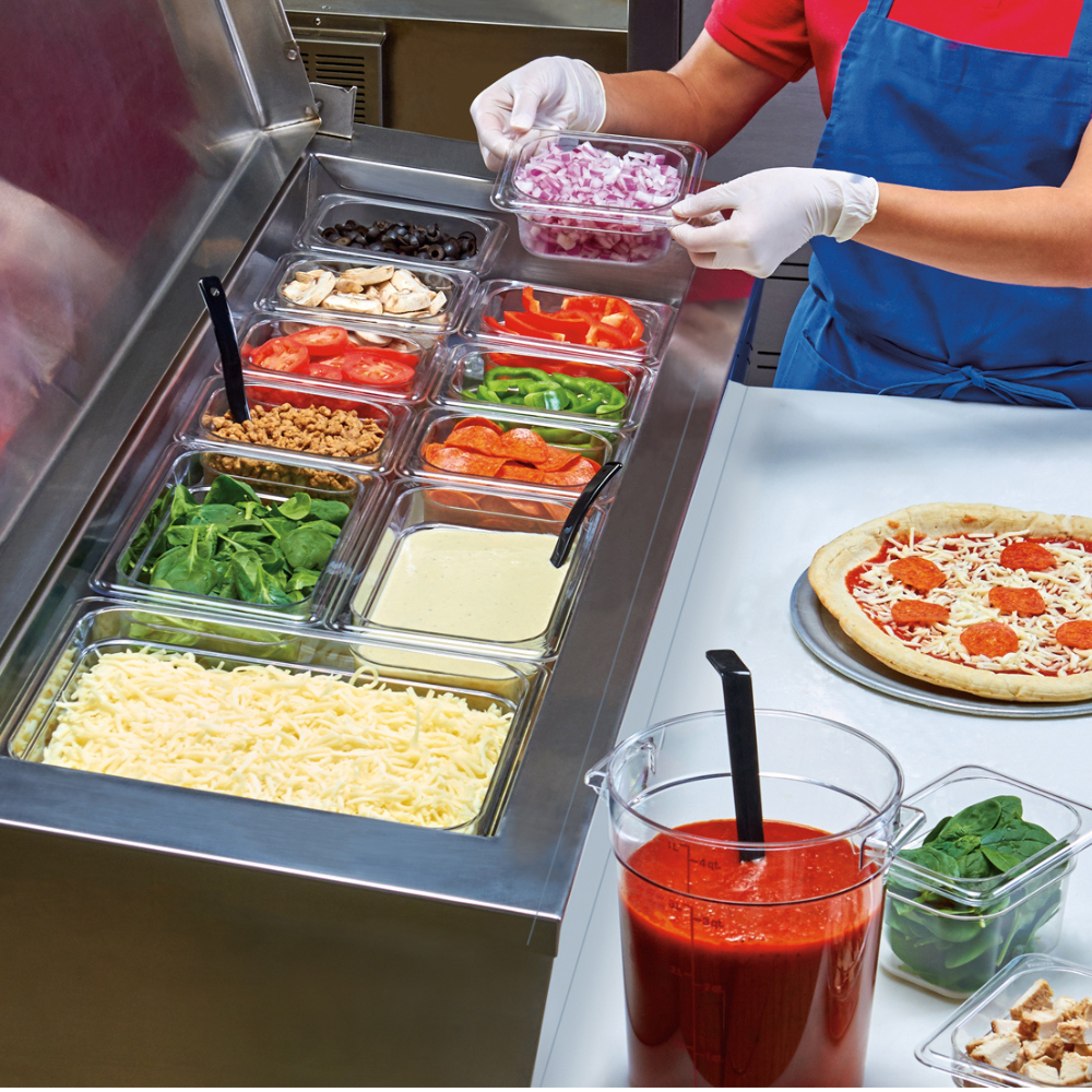 Cambro food pans in a pizza prep table next to a person preparing a pizza