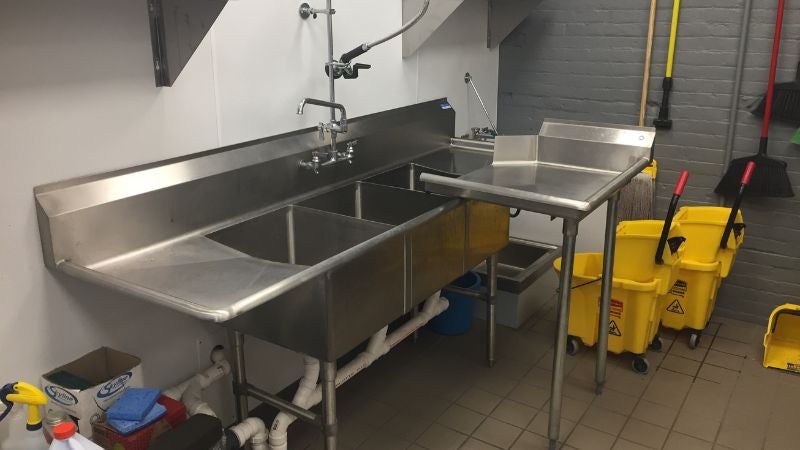 Commercial kitchen dishwashing station with triple sink, prep table, and cleaning supplies in culinary facility