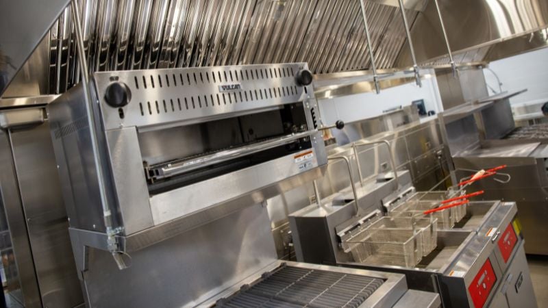 Commercial kitchen cookline with salamander broiler, fryers, and ventilation hood in school cafeteria kitchen