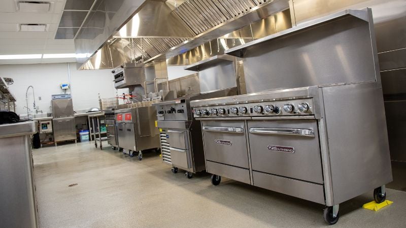 Commercial kitchen line with gas ranges, ovens, and stainless steel ventilation hood in school cafeteria kitchen