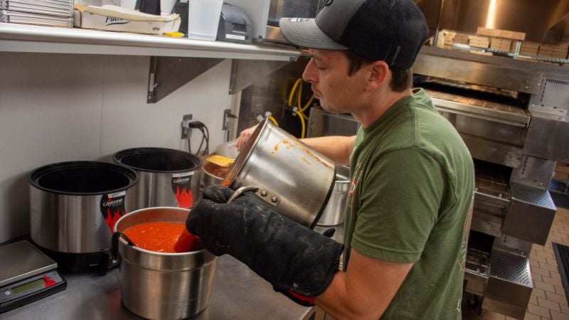 Staff transferring hot pizza sauce into a warming pot at prep station