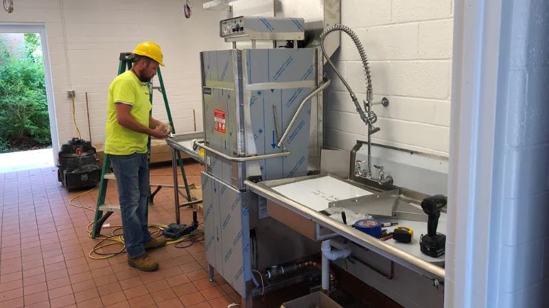 Worker in a hard hat installing a commercial dish machine next to a stainless steel sink, with tools and ladder in a kitchen workspace