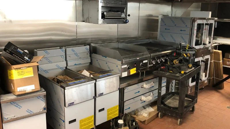 Commercial kitchen cooking line with deep fryers, flat-top griddle, and ovens, along with boxes and tools staged nearby during setup or installation