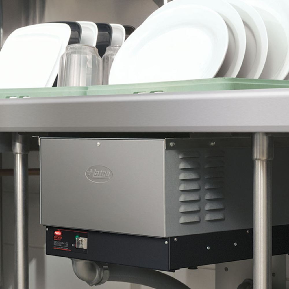 A stainless steel Hatco booster heater installed under a commercial dish table, positioned below a dish rack filled with clean white plates and clear glasses.