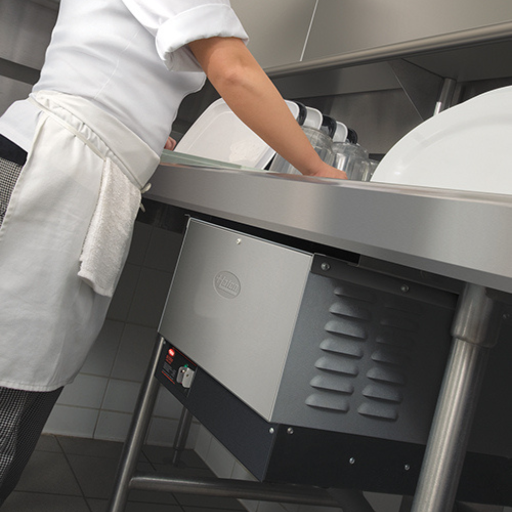 Kitchen worker unloading clean dishes from a stainless steel dish table with a Hatco booster heater installed below.