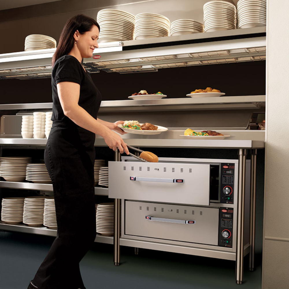 Kitchen worker using tongs to pull warm bread rolls from a Hatco stainless steel commercial drawer warmer.