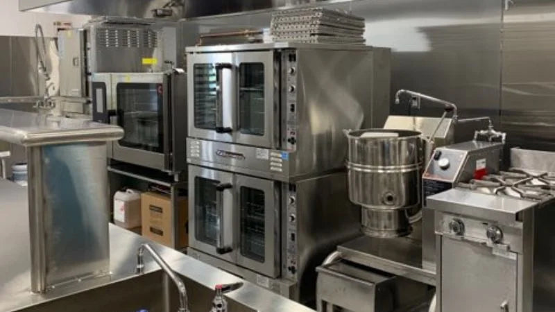 Commercial kitchen line with stacked convection ovens, large mixer, stock pot kettle, and gas range along stainless steel wall