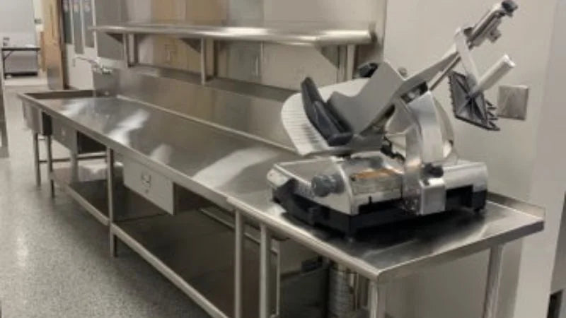 Stainless steel prep table with mounted shelving and a commercial meat slicer in a kitchen prep area