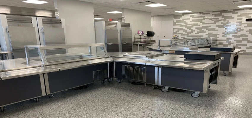 Cafeteria serving area with multiple stainless steel serving counters, sneeze guards, and refrigerators in the background