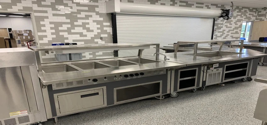 Stainless steel cafeteria serving line with hot wells, undercounter storage, and sneeze guard in front of a roll-up service door