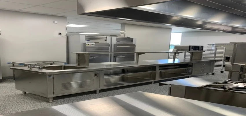 Large stainless steel kitchen prep and serving island with overhead ventilation hood and refrigerators in the background