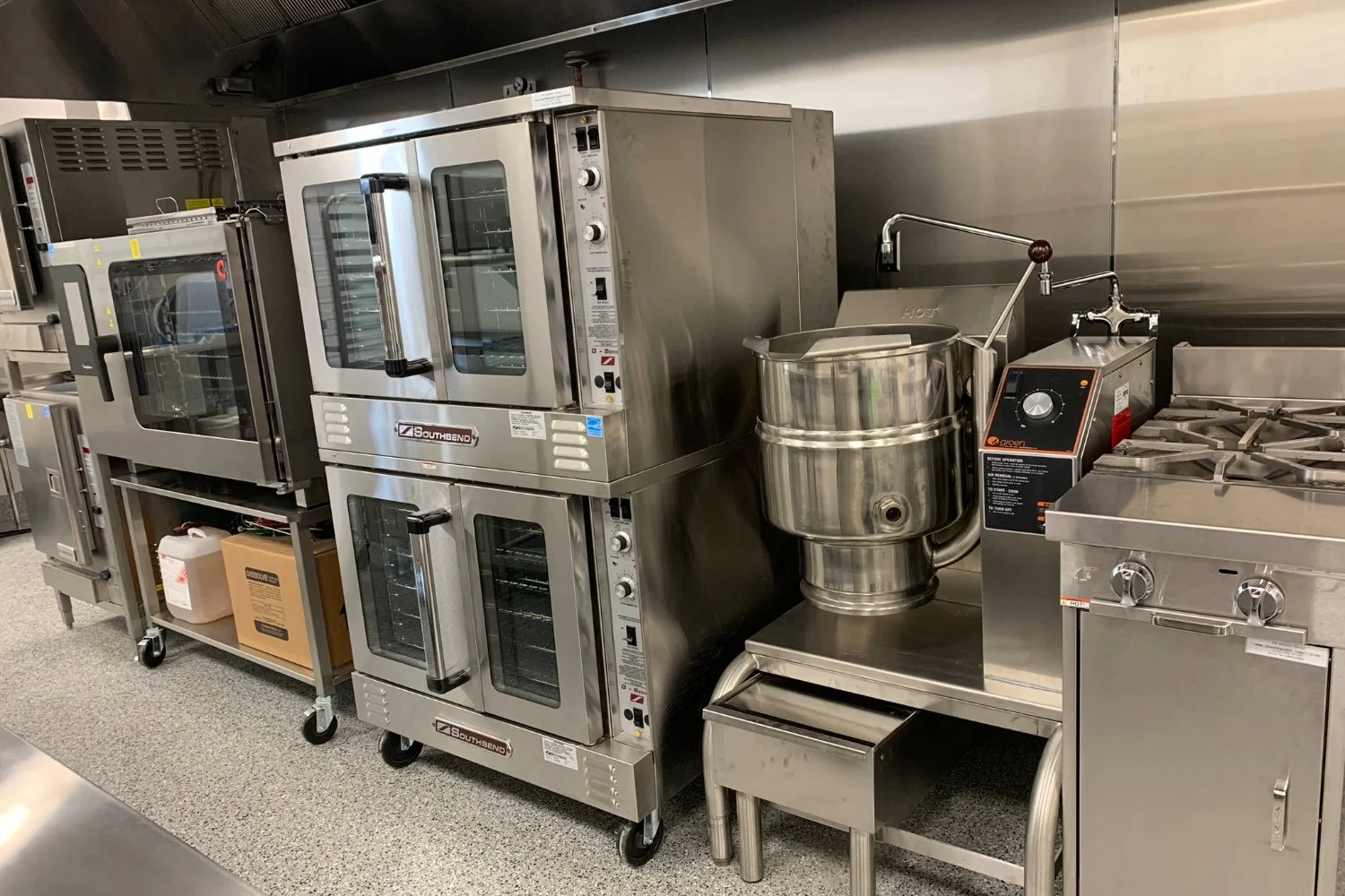 Commercial kitchen line with stacked convection ovens, large mixer, stock pot kettle, and gas range along stainless steel wall