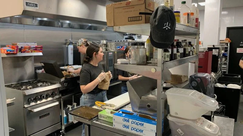 Busy commercial kitchen with staff preparing food at a cooking line, surrounded by shelves, ingredients, and stainless steel equipment