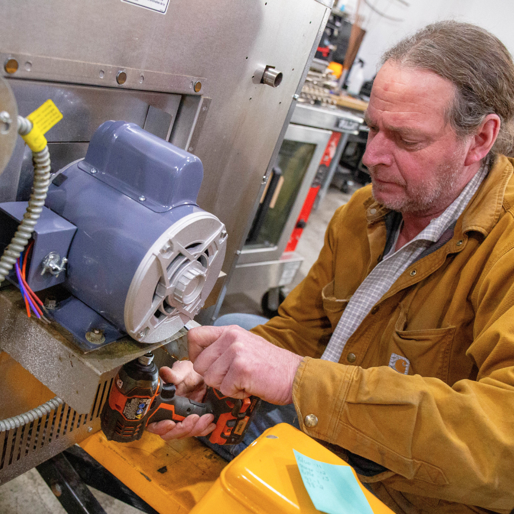 A technician using a power drill to repair a commercial motor on a stainless steel restaurant appliance.